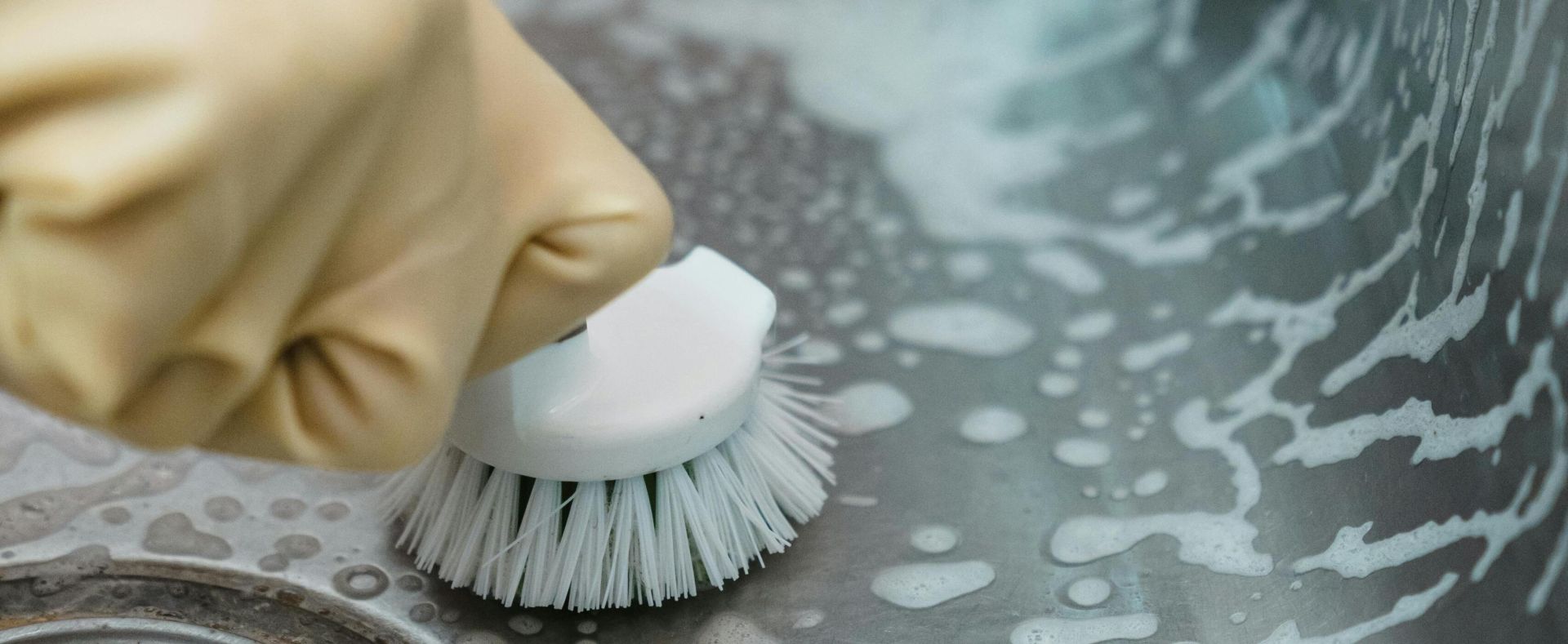 Close-up of a hand in gloves scrubbing a soapy kitchen sink with a brush.
